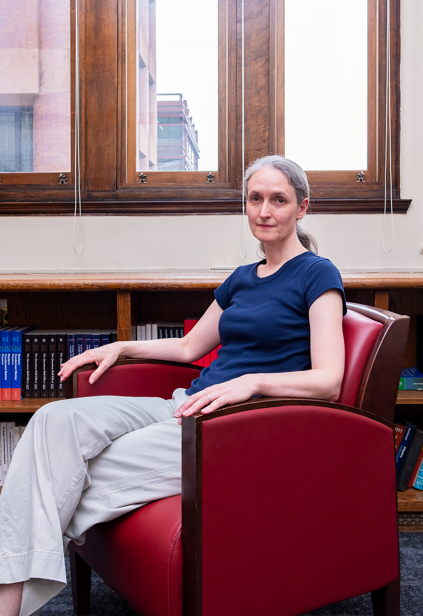 Demers sits in a burgundy chair in front of a window and a bookshelf.