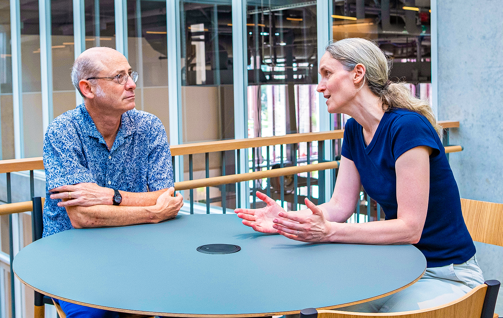 Demers sits at a table, conversing with a man wearing glasses.