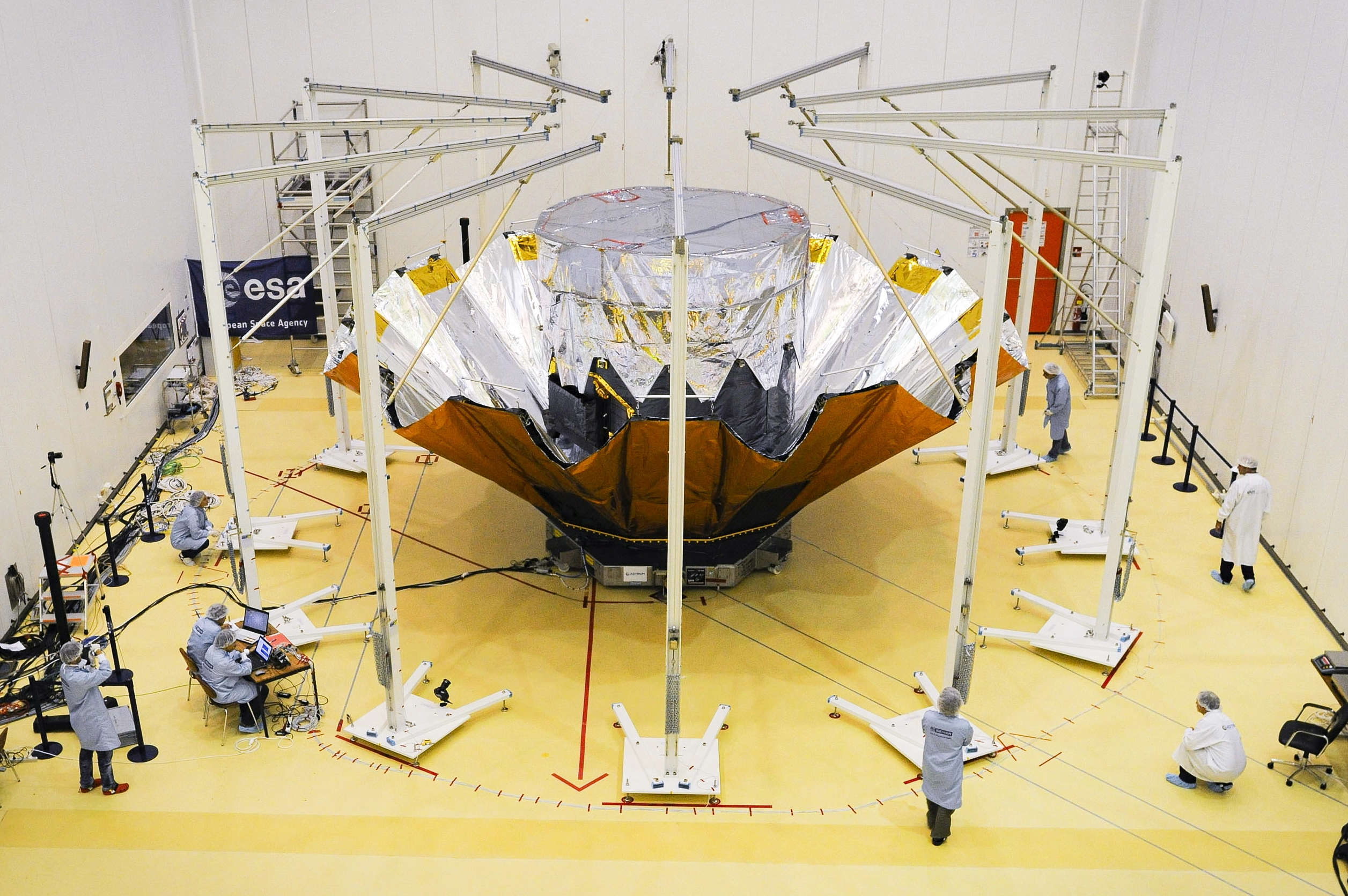 In a room with a yellow floor, eight scientists in lab coats and hairnets watch as a sunshield unfolds around a cylindrical spacecraft.