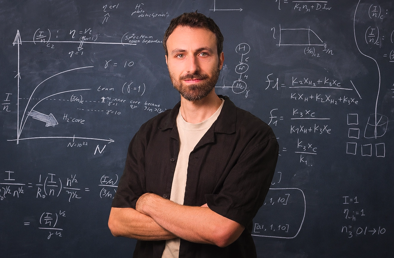 A man with a beard stands in front of a blackboard full of equations
