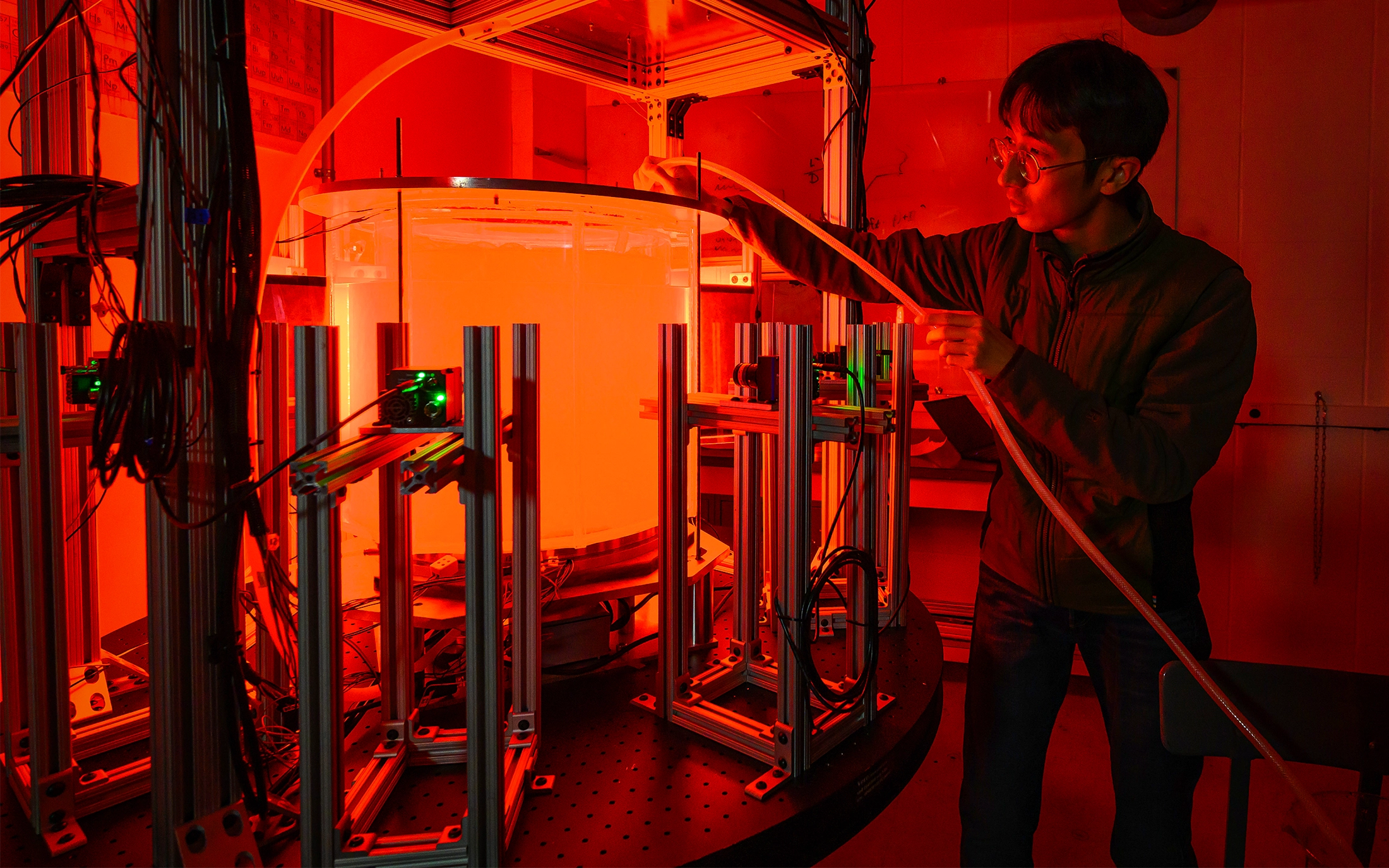 In a laboratory lit with red light, a man holds a tube over a tank filled with cloudy liquid.