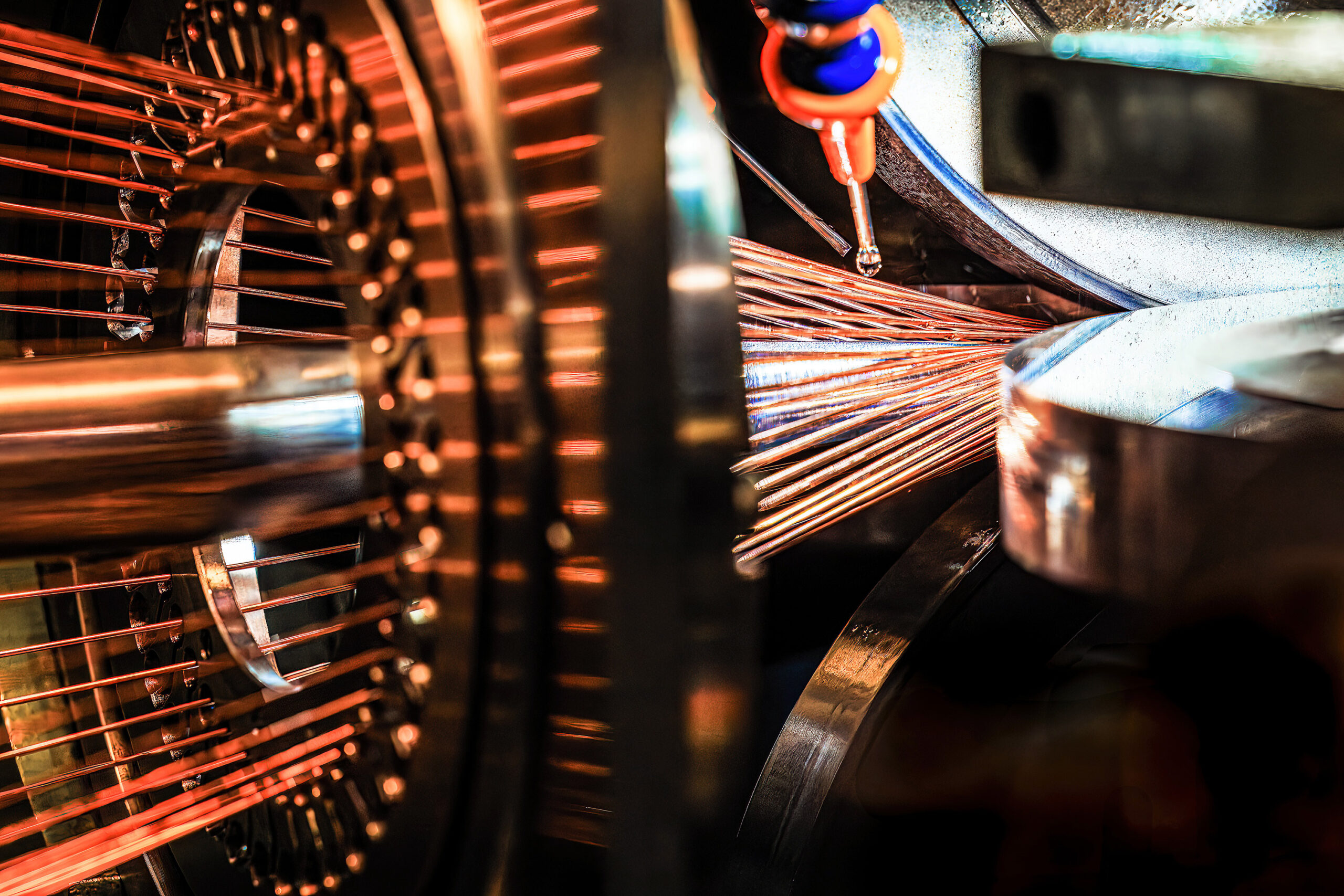 Close-up of the interior of a scientific machine. On the left, a circular arrangement of evenly spaced copper-colored rods radiates outward from a central hub. The bundle of thin copper wires converges on the right into a silver cylindrical component.