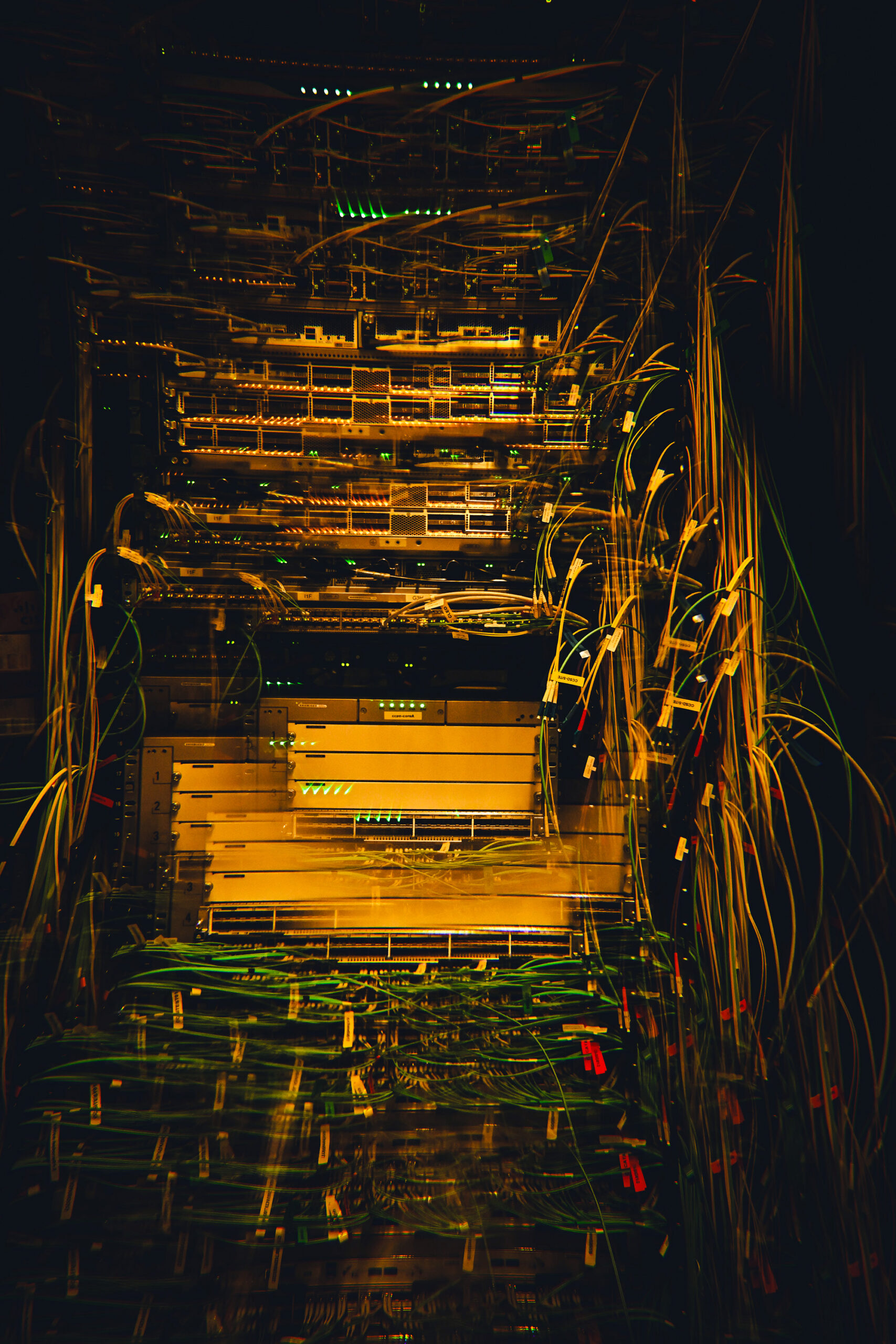 Rows of densely cabled server racks in a darkened data center. Hundreds of yellow, green, and white cables run loosely down the sides of the racks in tangled bundles.
