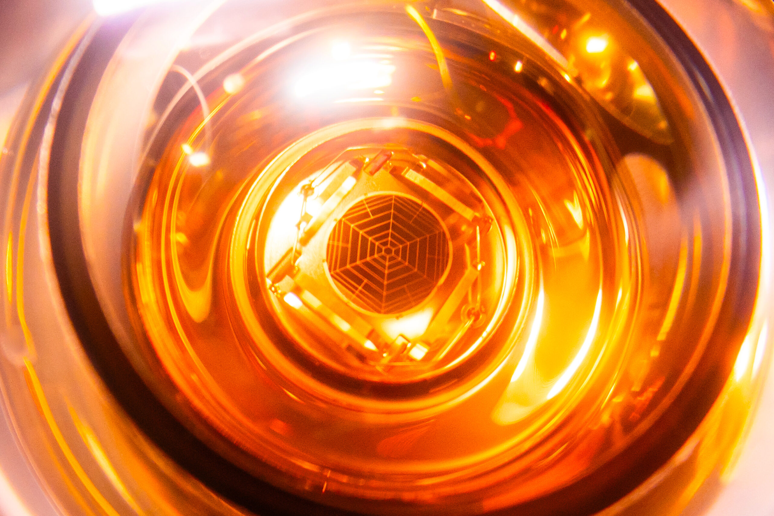 Close-up photograph looking into a curved sensor device in warm golden light. At the center, a small geometric spider web-like grid pattern is visible.