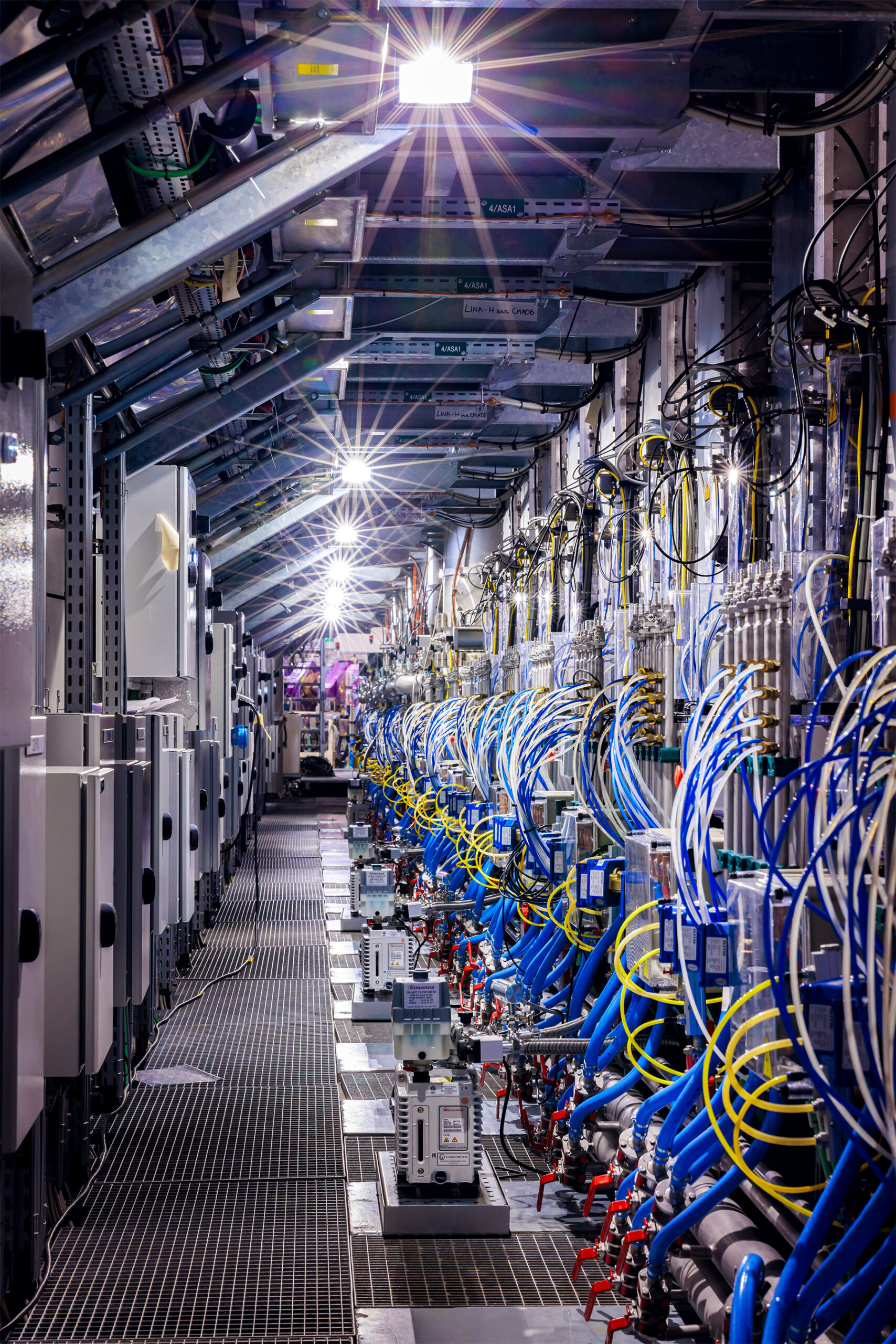 A long, narrow industrial corridor stretching into the distance, lined on both sides with densely packed machinery, pipes, and bundles of blue and yellow cables. Bright overhead lights illuminate the passage, and the floor is made of metal grating.