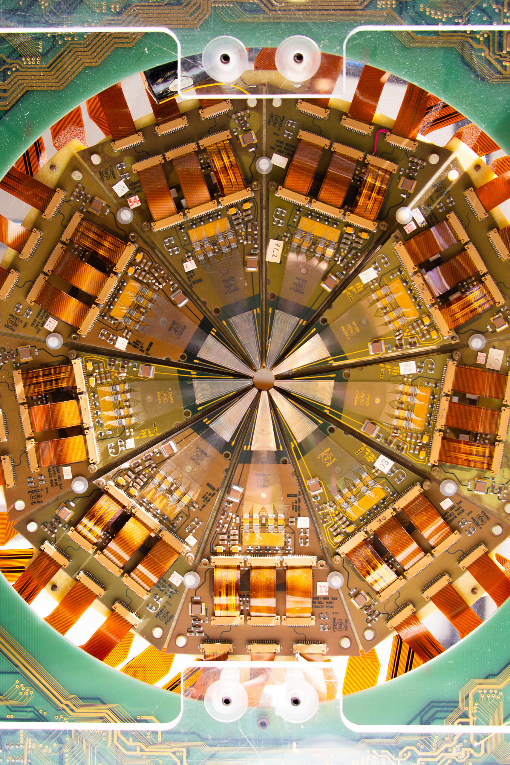 A circular scientific instrument with a radially symmetric layout. The interior is divided into repeating wedge-shaped segments arranged around a central hub, each containing densely packed electronic circuit boards, copper-colored wiring, and gold and orange components.