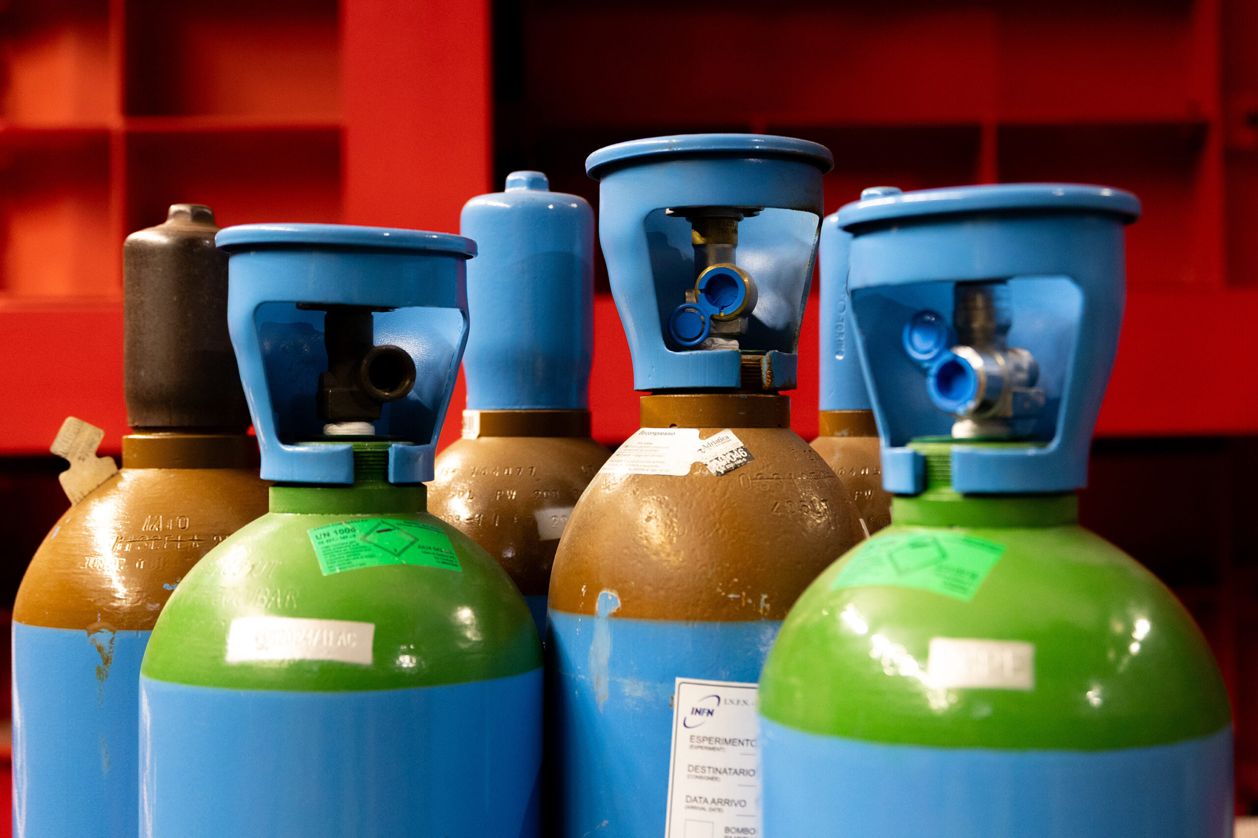 Close-up of six industrial gas cylinders grouped closely together against a bright red background. Two cylinders have green tops, four have brown tops. The green cylinders and one of the brown cylinders have blue valve caps attached at their tops.