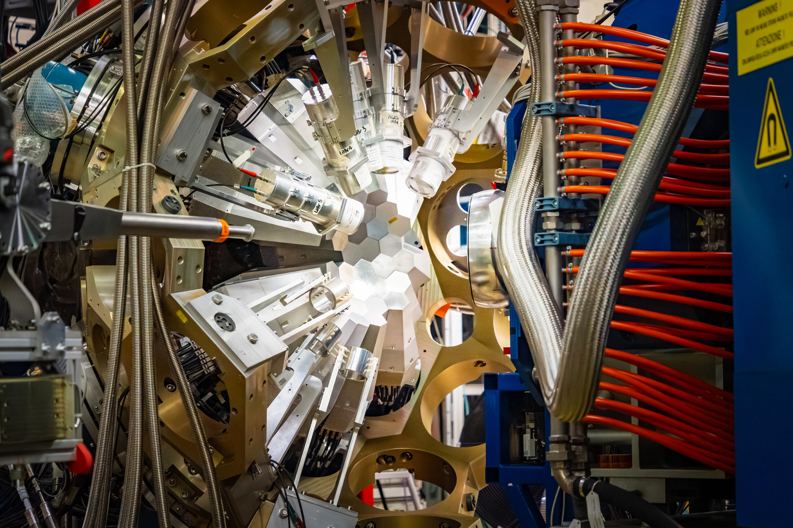 A large, complex scientific machine with a symmetrical, circular arrangement of white hexagonal panels at its center, surrounded by gold-colored metal arms. Numerous cables, flexible metallic hoses, and bright orange tubes extend from the structure in multiple directions.