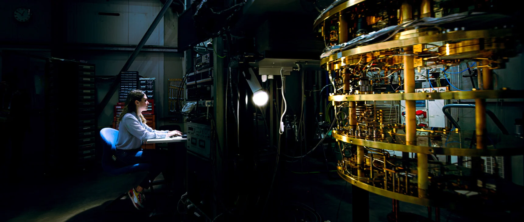 A researcher sits at a keyboard at a workstation illuminated by a single light in a darkened lab facility. To the right is a large piece of complex machinery featuring stacked rings of gold-colored metal components and intricate circuitry.