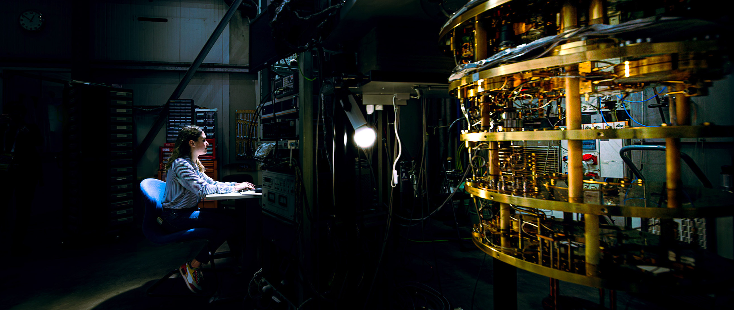 A researcher sits at a keyboard at a workstation illuminated by a single light in a darkened lab facility. To the right is a large piece of complex machinery featuring stacked rings of gold-colored metal components and intricate circuitry.