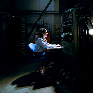 A researcher sits at a keyboard at a workstation illuminated by a single light in a darkened lab facility. To the right is a large piece of complex machinery featuring stacked rings of gold-colored metal components and intricate circuitry.