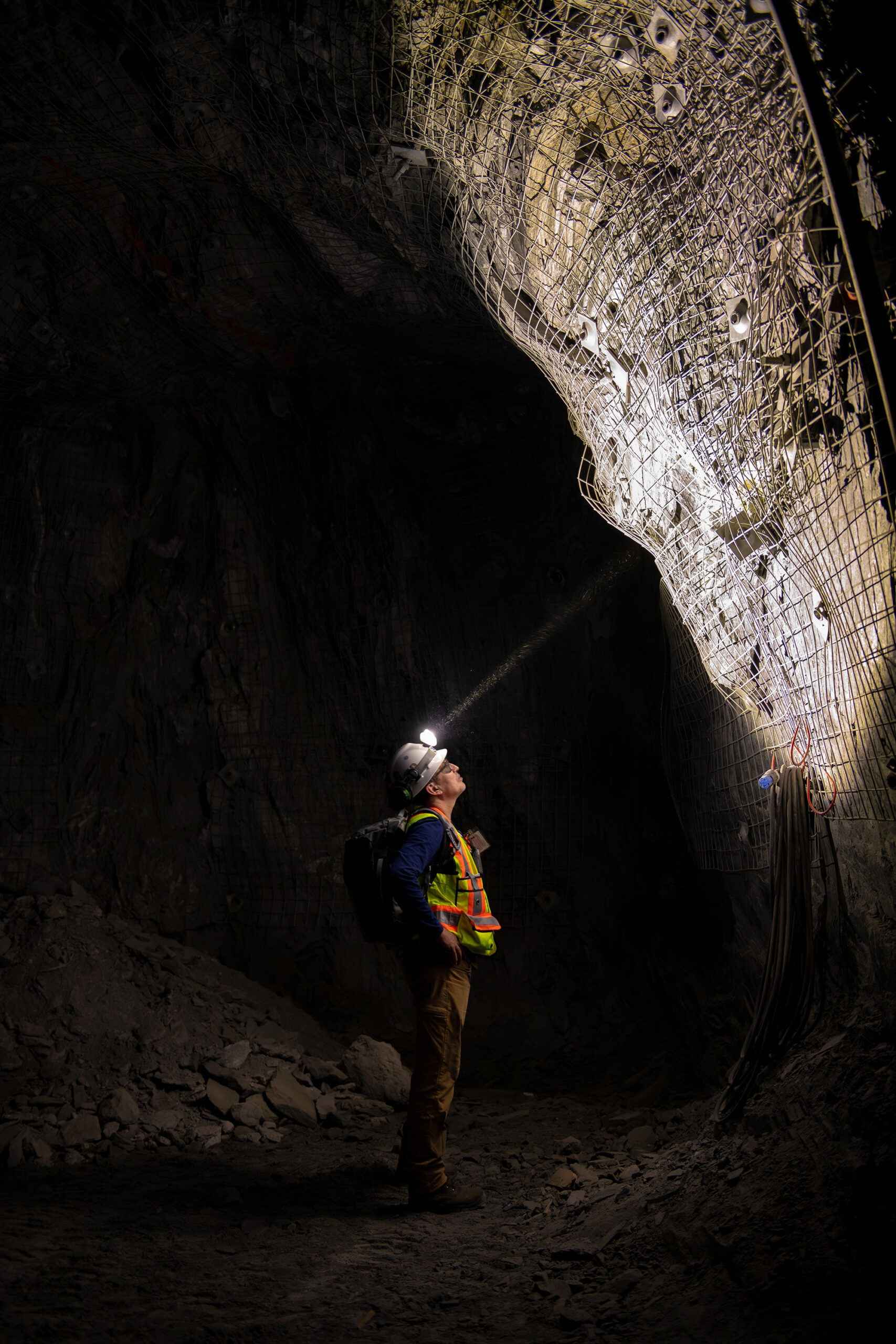 A person wearing a hard hat with a headlamp, a high-visibility safety vest, and a backpack stands inside a large, dark underground tunnel looking upward. The headlamp beam illuminates the walls above and to his right.
