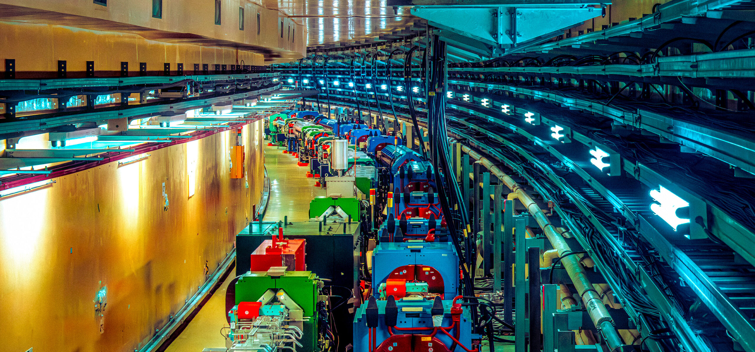 A view along a long, curving underground tunnel housing a series of large scientific machines. The wall on the left has golden-yellow paneling. On the right, multiple black cables run along the tunnel wall in parallel.