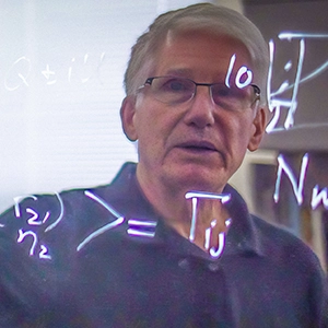 A man with white hair and glasses is seen reflected in a shiny blackboard.