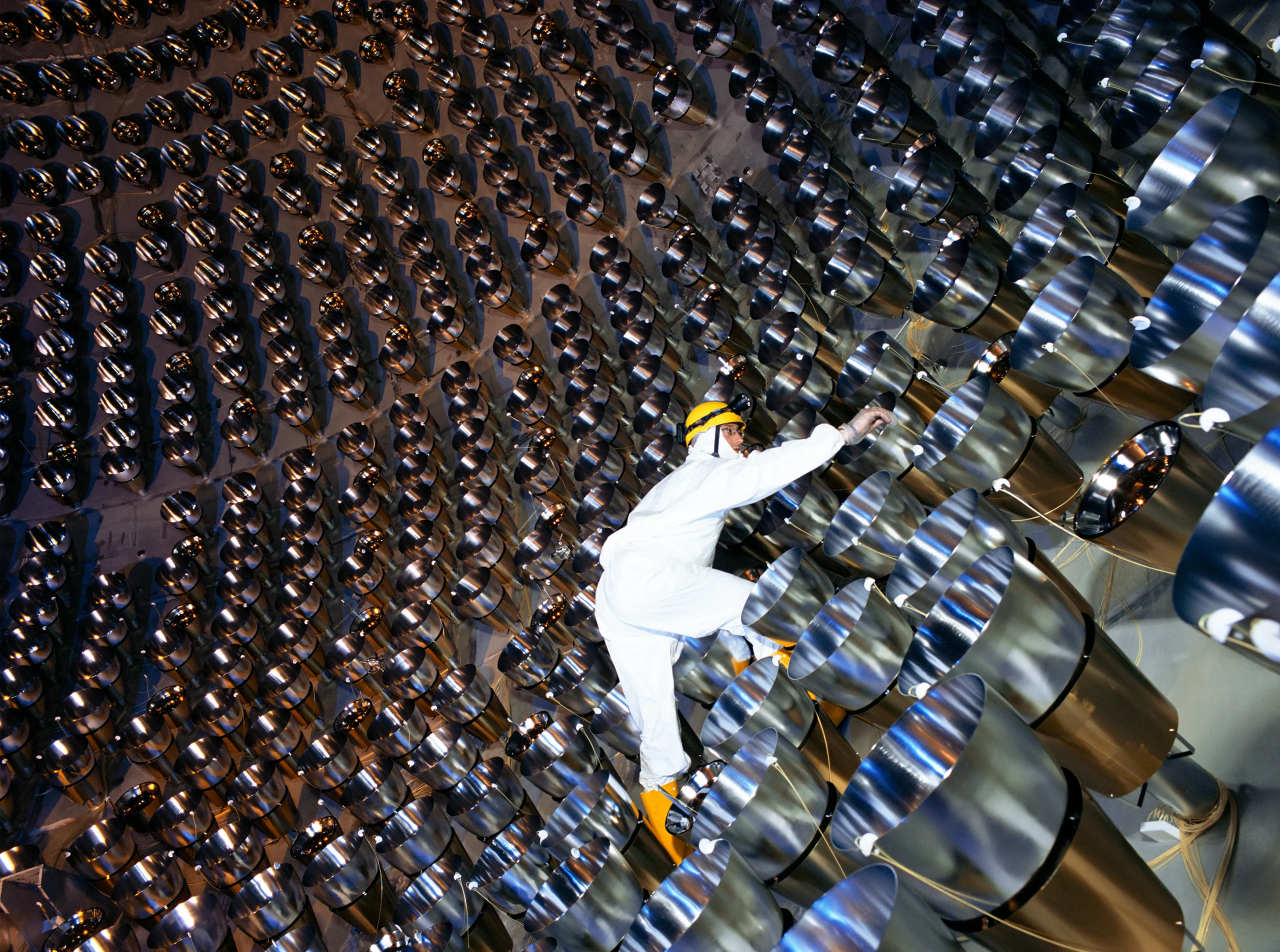A technician in a white cleanroom suit and yellow hard hat crawls across the curved inner surface of a neutrino detector lined with hundreds of inward-facing photomultiplier tubes.