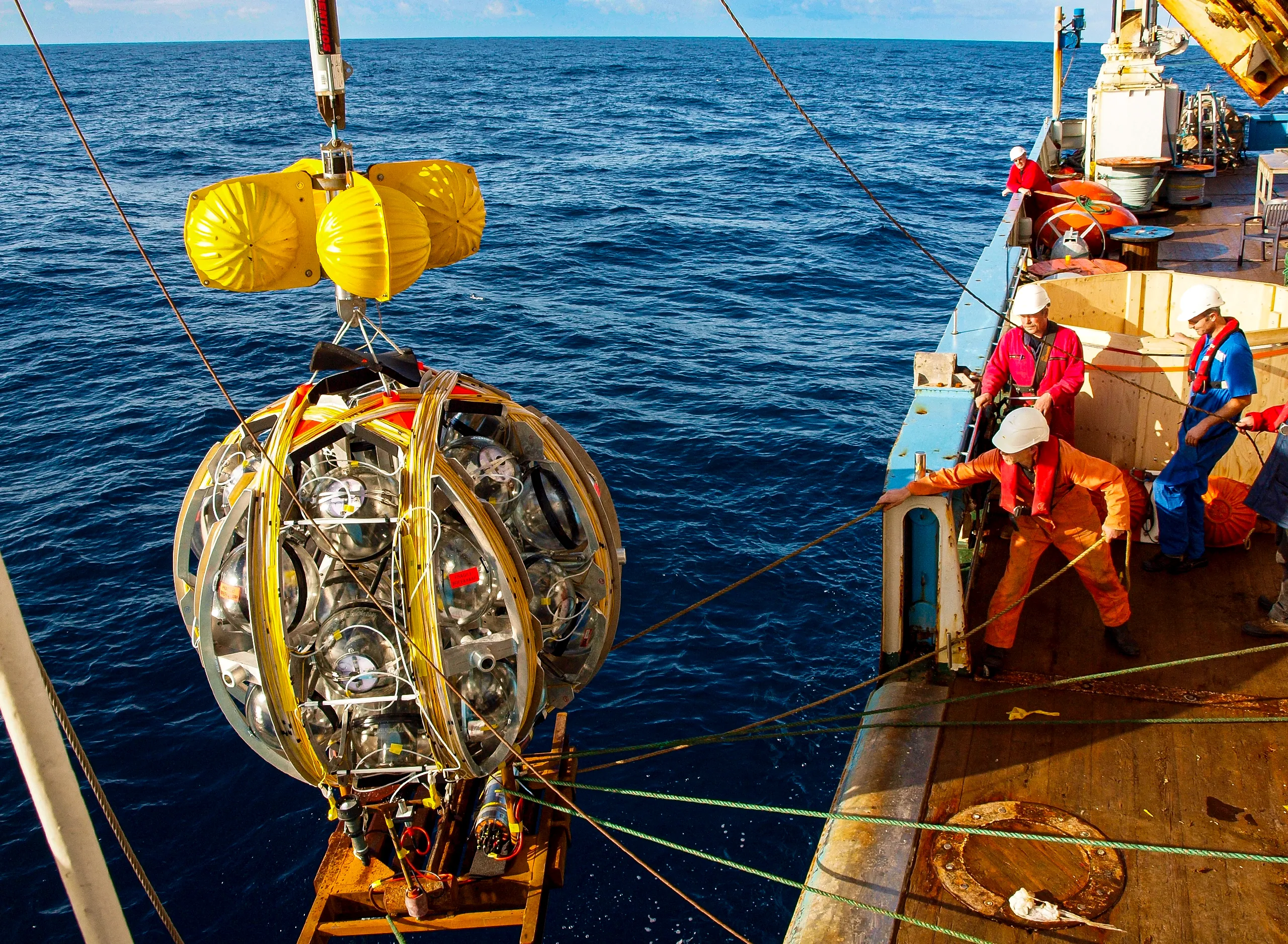 Crew members on a research vessel deploy a spherical detector module using a crane over open ocean, with yellow buoyancy floats attached on top.
