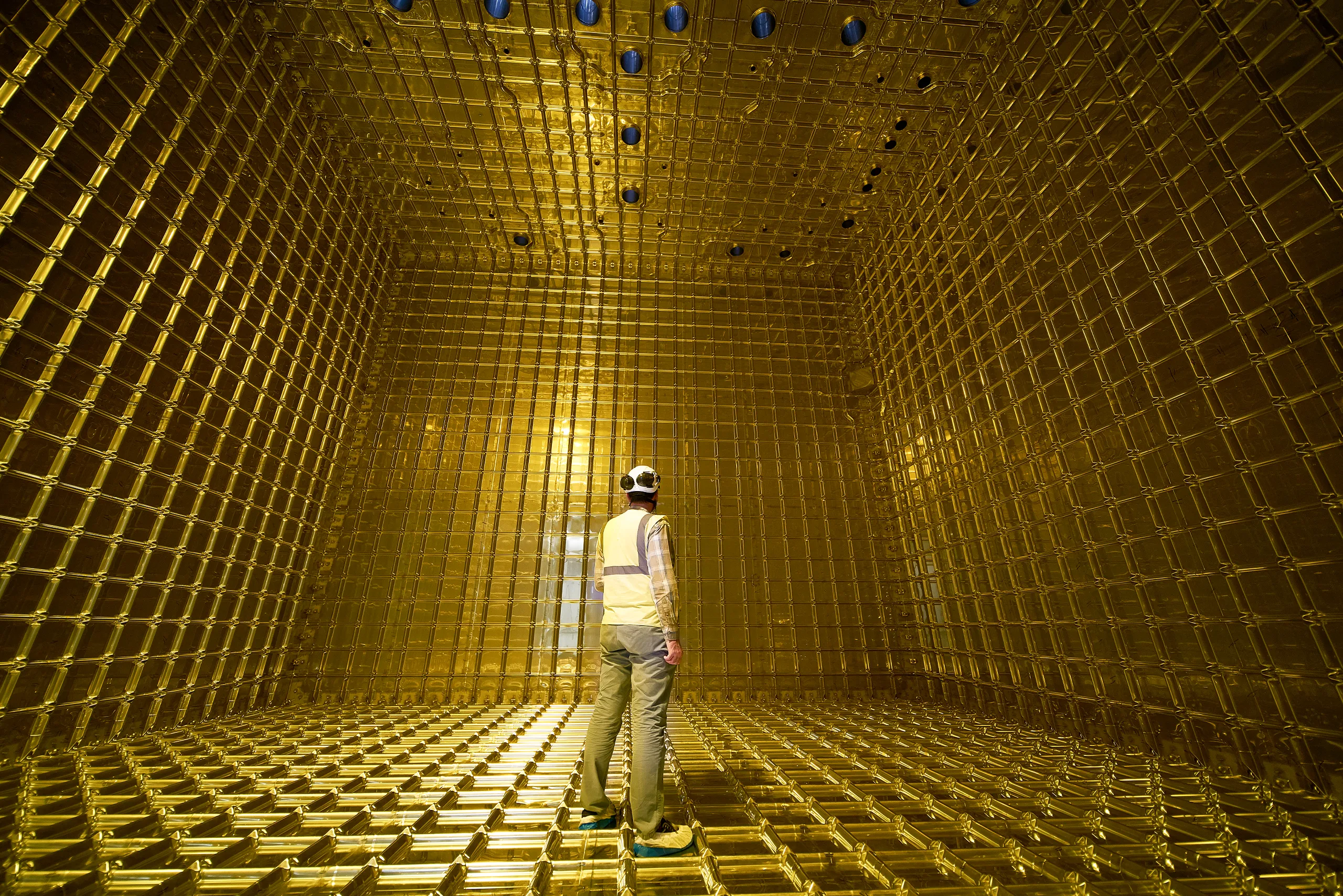 A worker in a hard hat and safety vest stands inside a large rectangular space lined entirely with a bright yellow wire-frame grid structure.