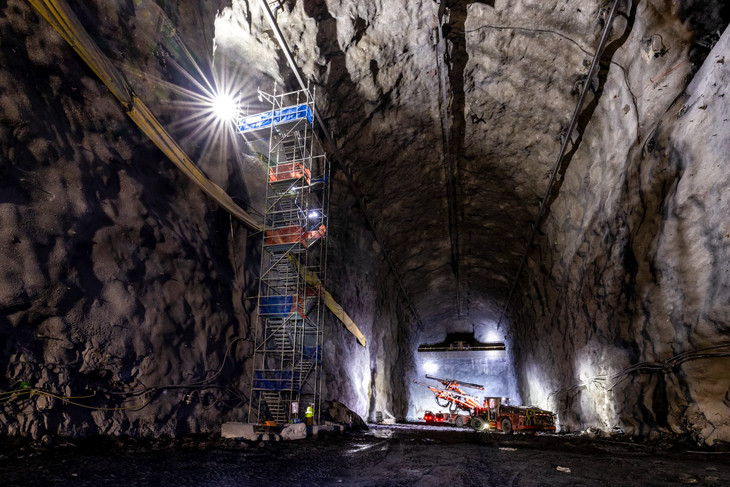 Underground excavation of a large rock cavern for a physics experiment, with scaffolding, bright lighting, and heavy drilling machinery visible inside the newly blasted tunnel.