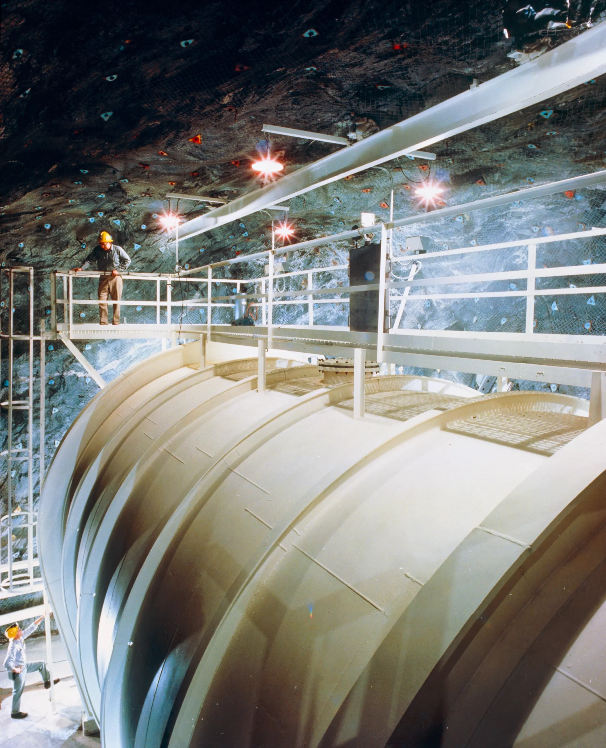 Workers in hard hats inspect a large industrial tank inside an underground rock cavern, with metal walkways and bright work lights overhead.