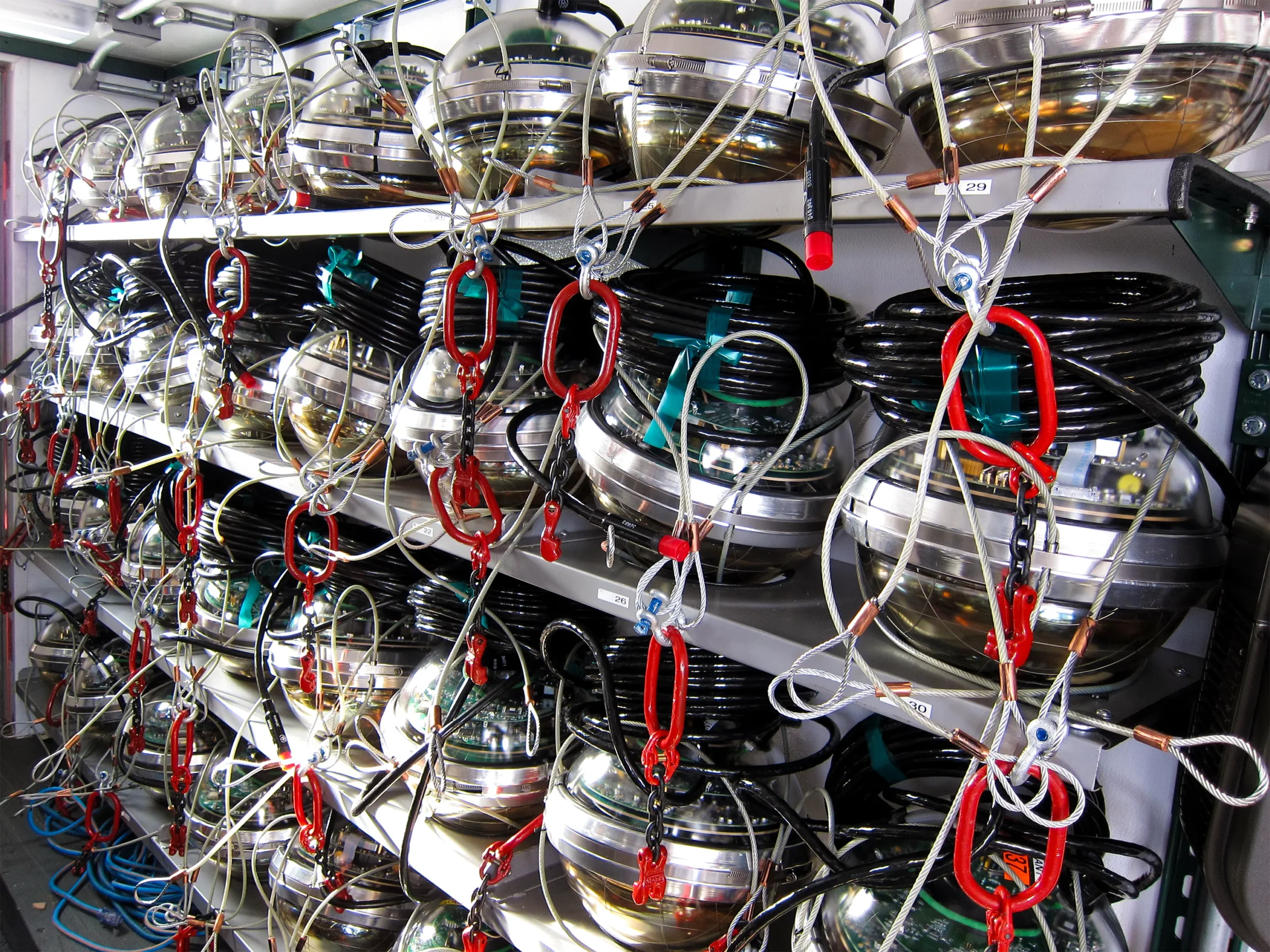 Shelves stacked with numerous spherical detectors in various stages of assembly, with wiring and red carabiner clips visible, in a storage or preparation area.
