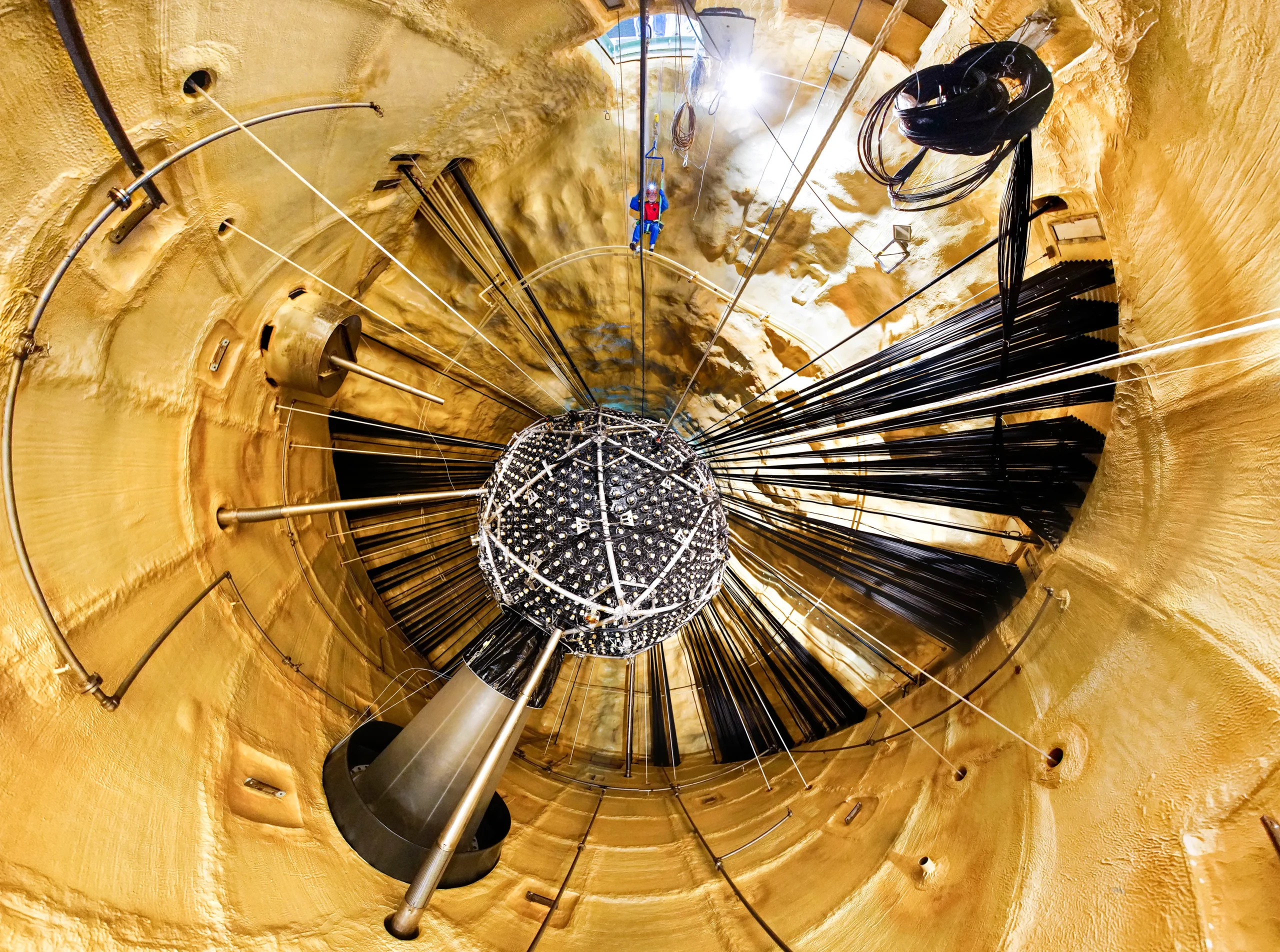 Wide-angle view showing a small central sphere surrounded by cables and support structures inside a cylindrical underground chamber. A man hangs from support structures.