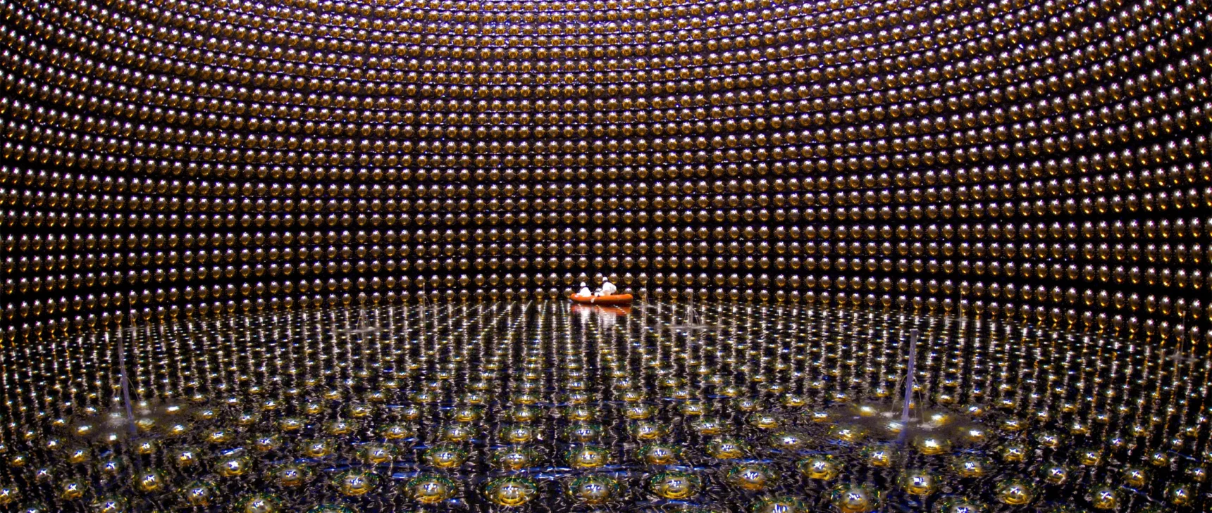 A large cavern lined with reflective spheres and partially filled with water. Two people in white protective gear float in an orange raft in the distance.