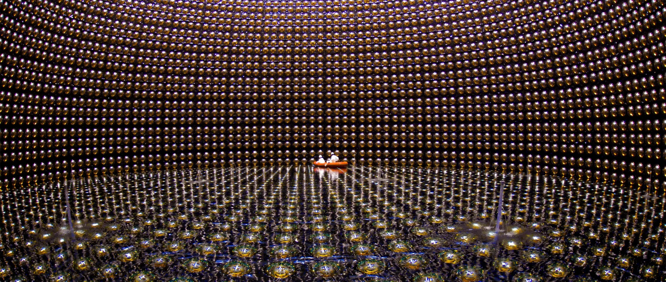 A large cavern lined with reflective spheres and partially filled with water. Two people in white protective gear float in an orange raft in the distance.