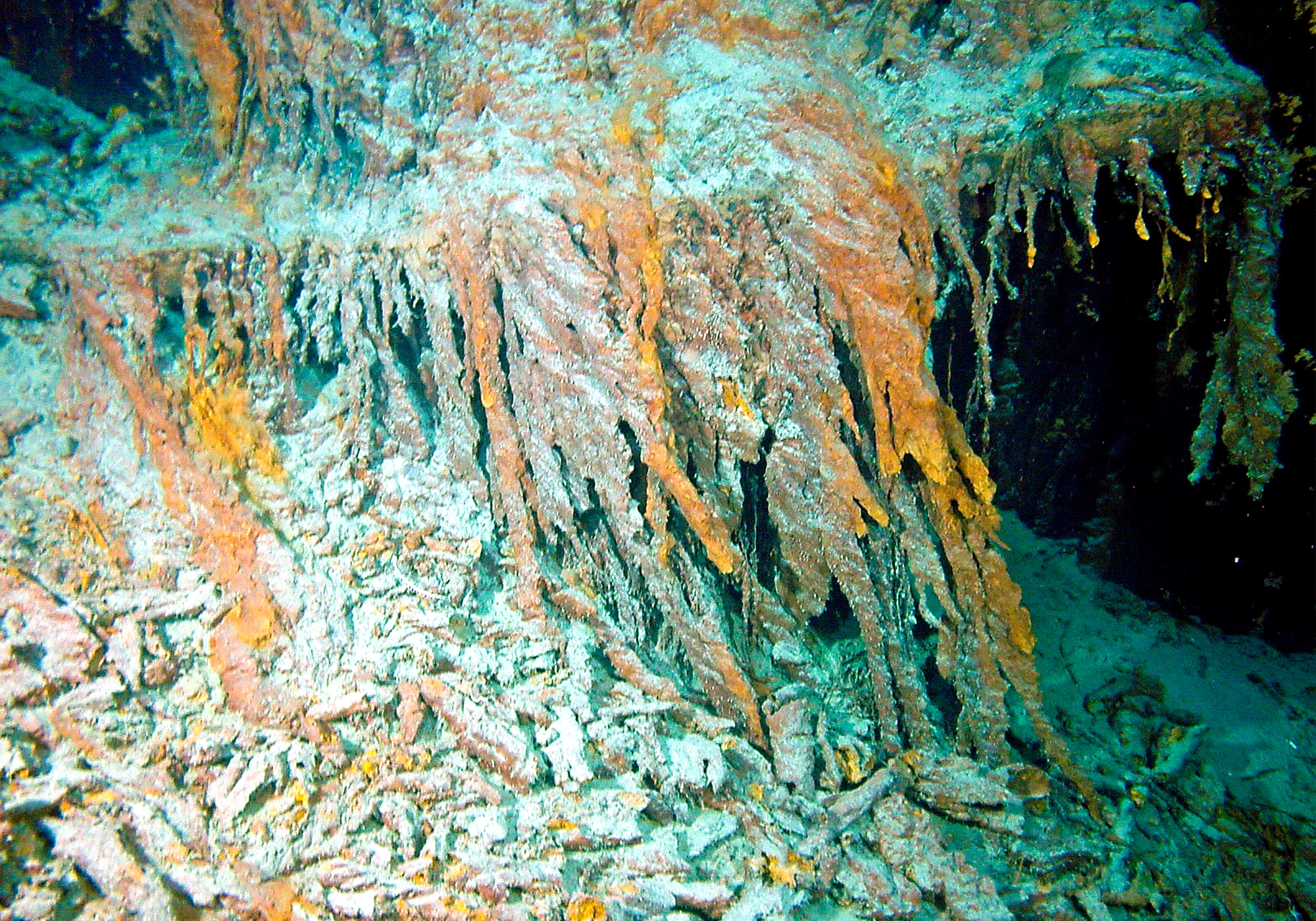 Close-up of rust colored, icicle-like mineral or rust formations hanging in clusters, with debris scattered on the seafloor.