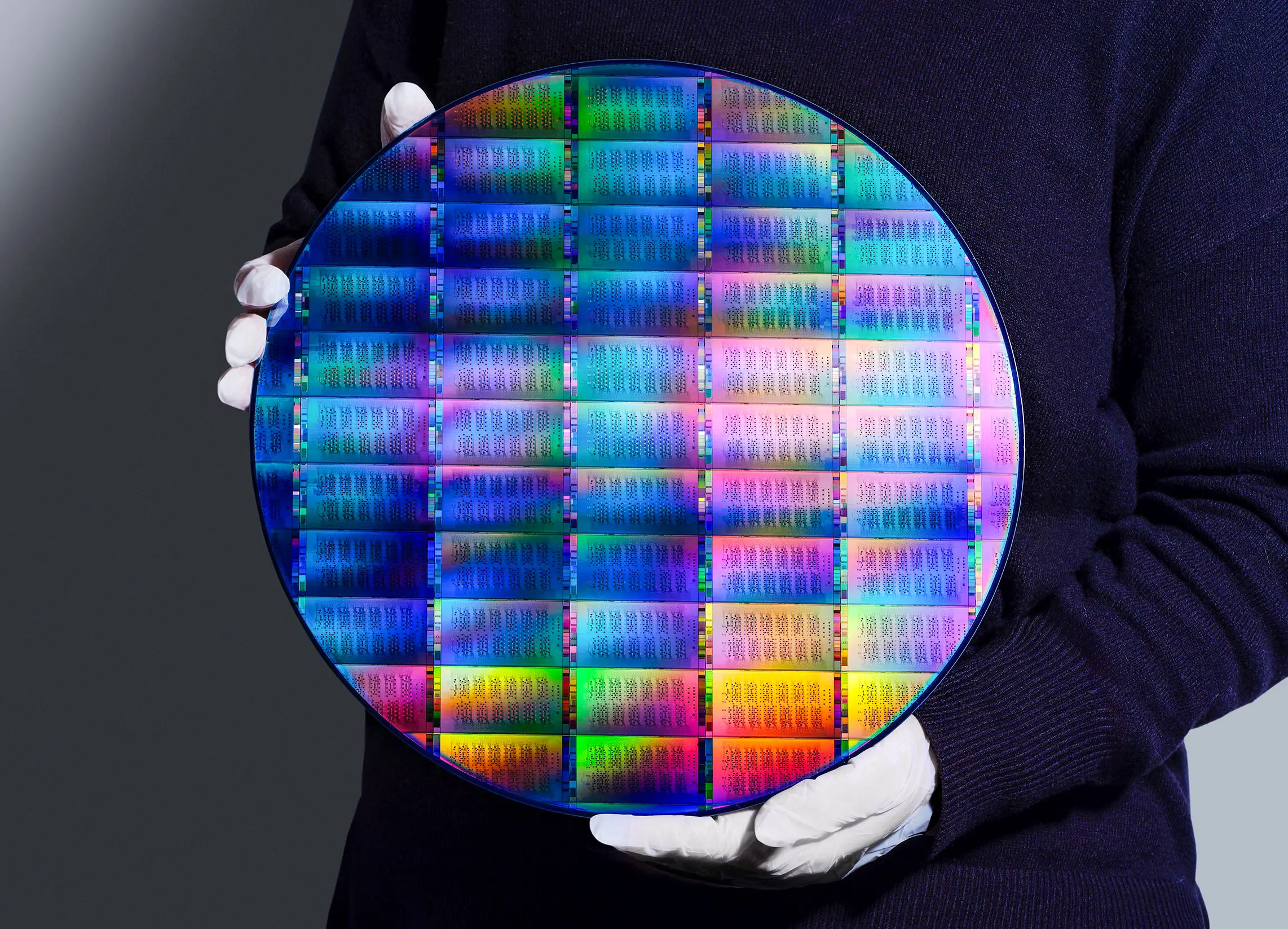 A person in white gloves holding a circular wafer etched with a grid of rectangular chip patterns. Rainbow colors shine across the wafer from light diffraction.