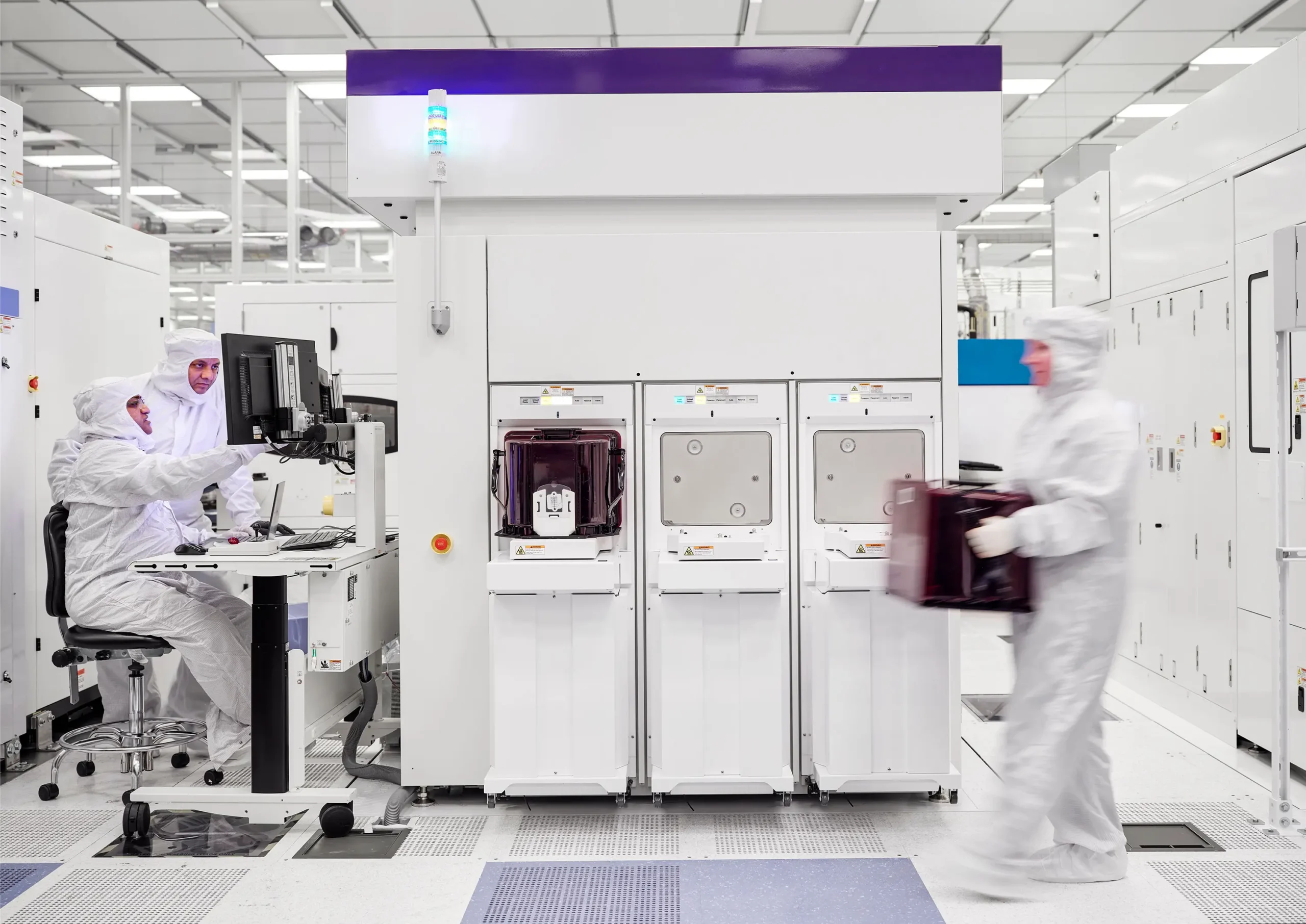 Three workers in white cleanroom suits operate semiconductor fabrication equipment. Two are seated at a computer terminal and is one carrying a box across the floor.