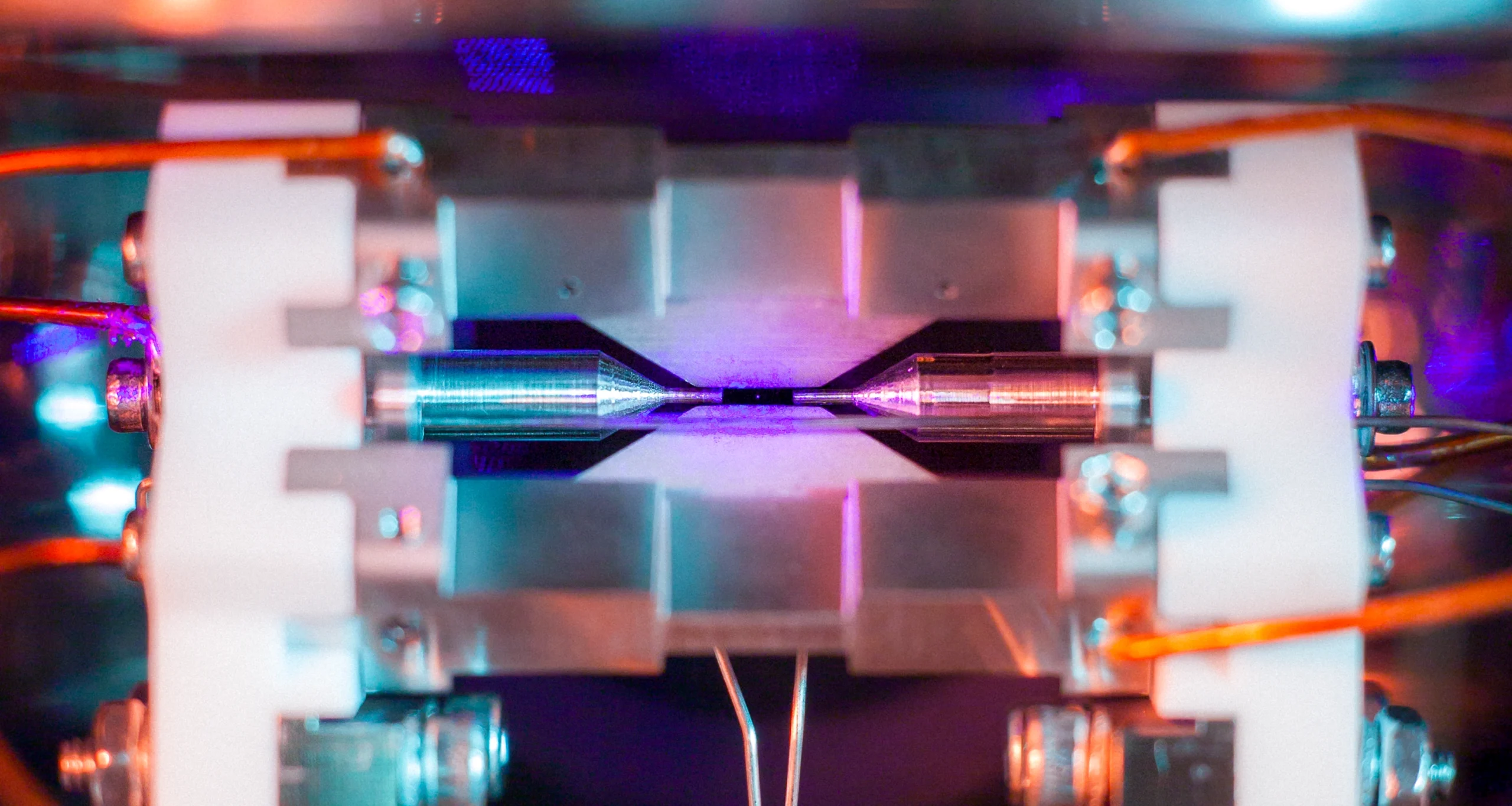 Close-up of a linear ion trap inside a vacuum chamber, showing metal electrodes and a glowing purple ion cloud held between two sharp needle-like tips.