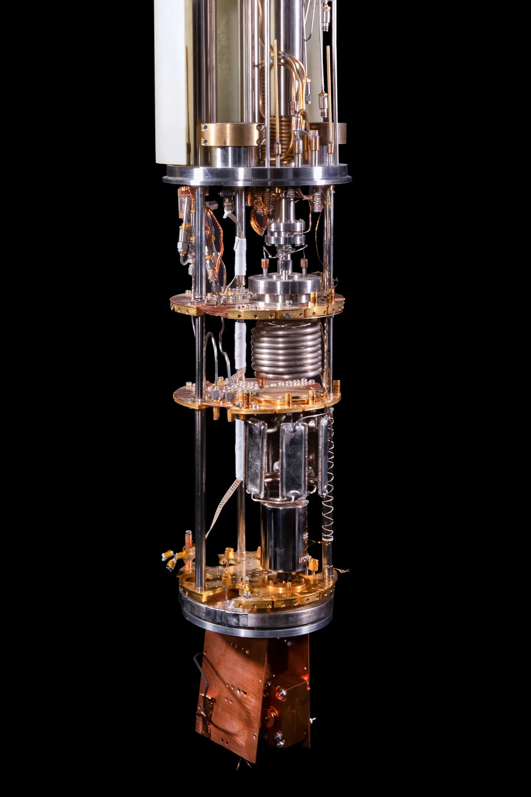A chandelier-like dilution refrigerator is suspended vertically, with multiple gold-colored circular plates connected by rods, wiring, and a coiled tube.
